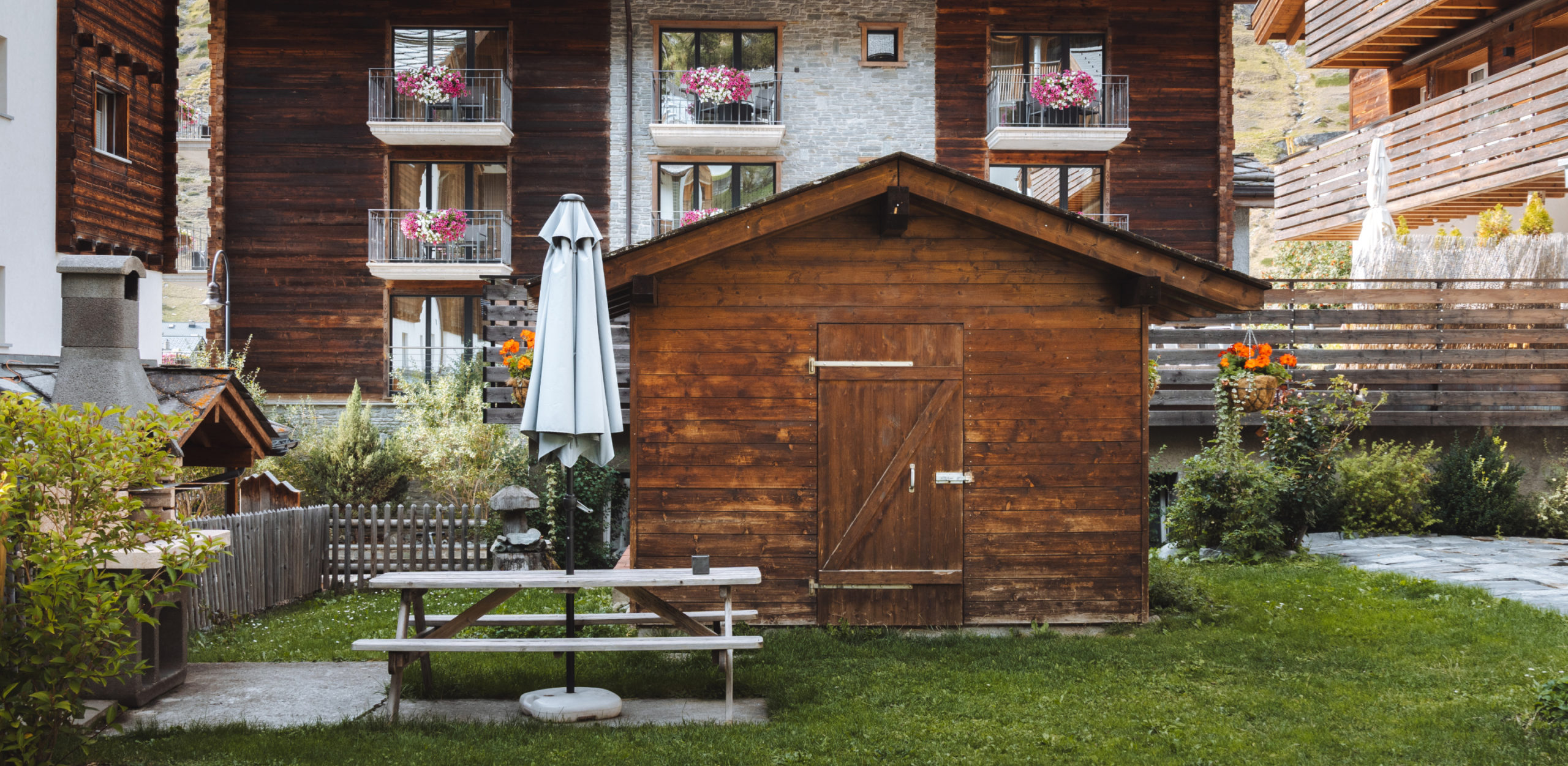 Petite maison en bois dans le jardin avec banc et chalets environnants en arrière-plan.