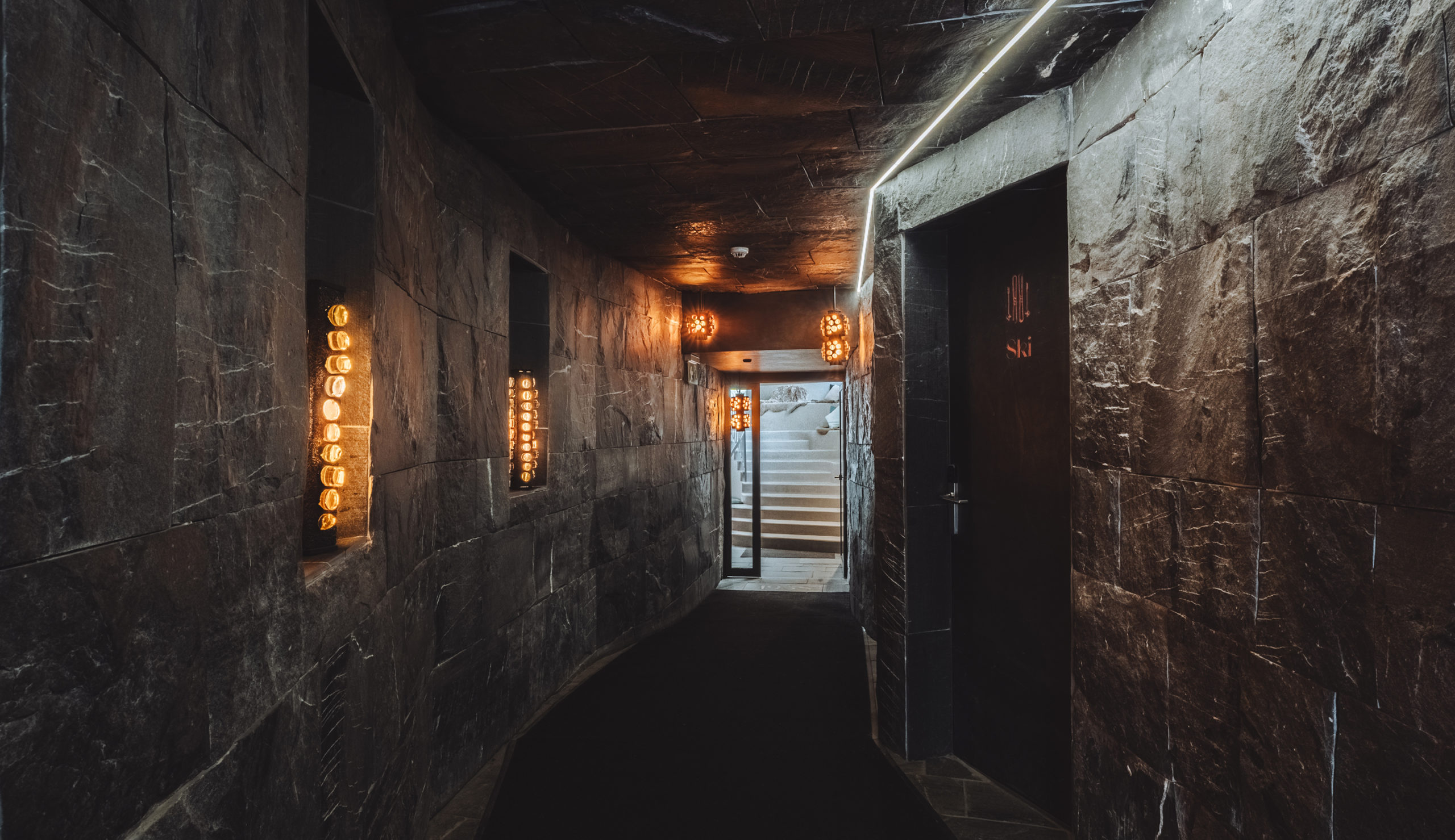 Couloir sombre aux murs en pierre et éclairage mural à l’Hôtel Carina Zermatt, avec un escalier au bout du passage