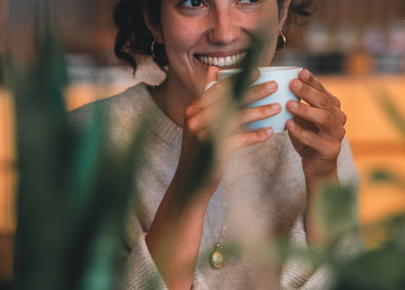 Frau lächelt und hält eine Tasse Heißgetränk in gemütlicher, warm beleuchteter Umgebung.