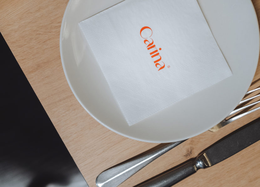 Teller mit Serviette und Carina-Logo neben Besteck auf einem Holztisch.