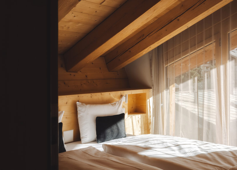 Chambre avec habillage en bois et lit double à côté d’une grande fenêtre avec voilage.