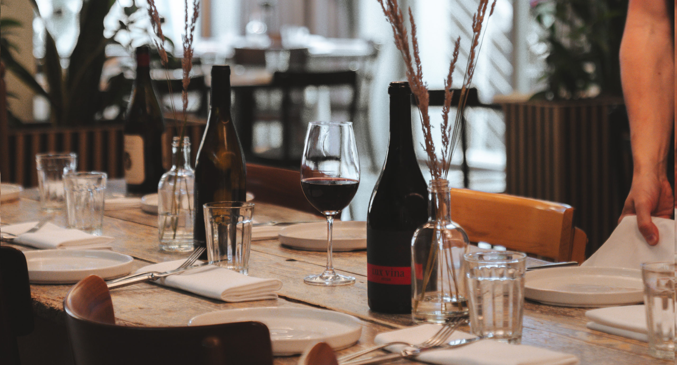 Gedeckter Tisch im Restaurant des Hotel Carina Zermatt mit Gläsern, Weinflaschen und Servietten; eine Hand legt eine Serviette auf