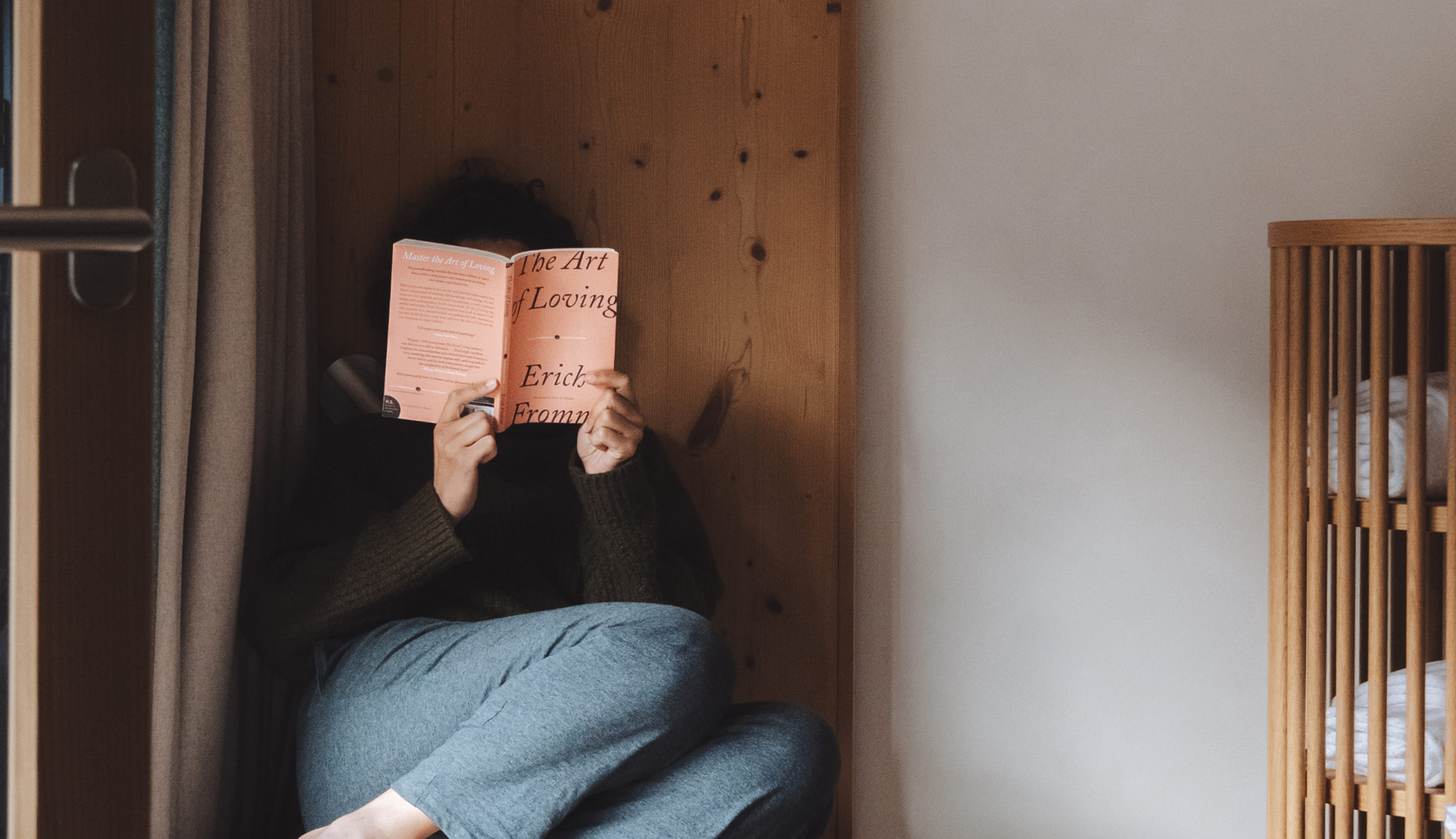 Personne assise dans un coin d’une chambre à l’Hôtel Carina Zermatt en train de lire un livre; une étagère en bois avec des serviettes pliées se trouve à côté